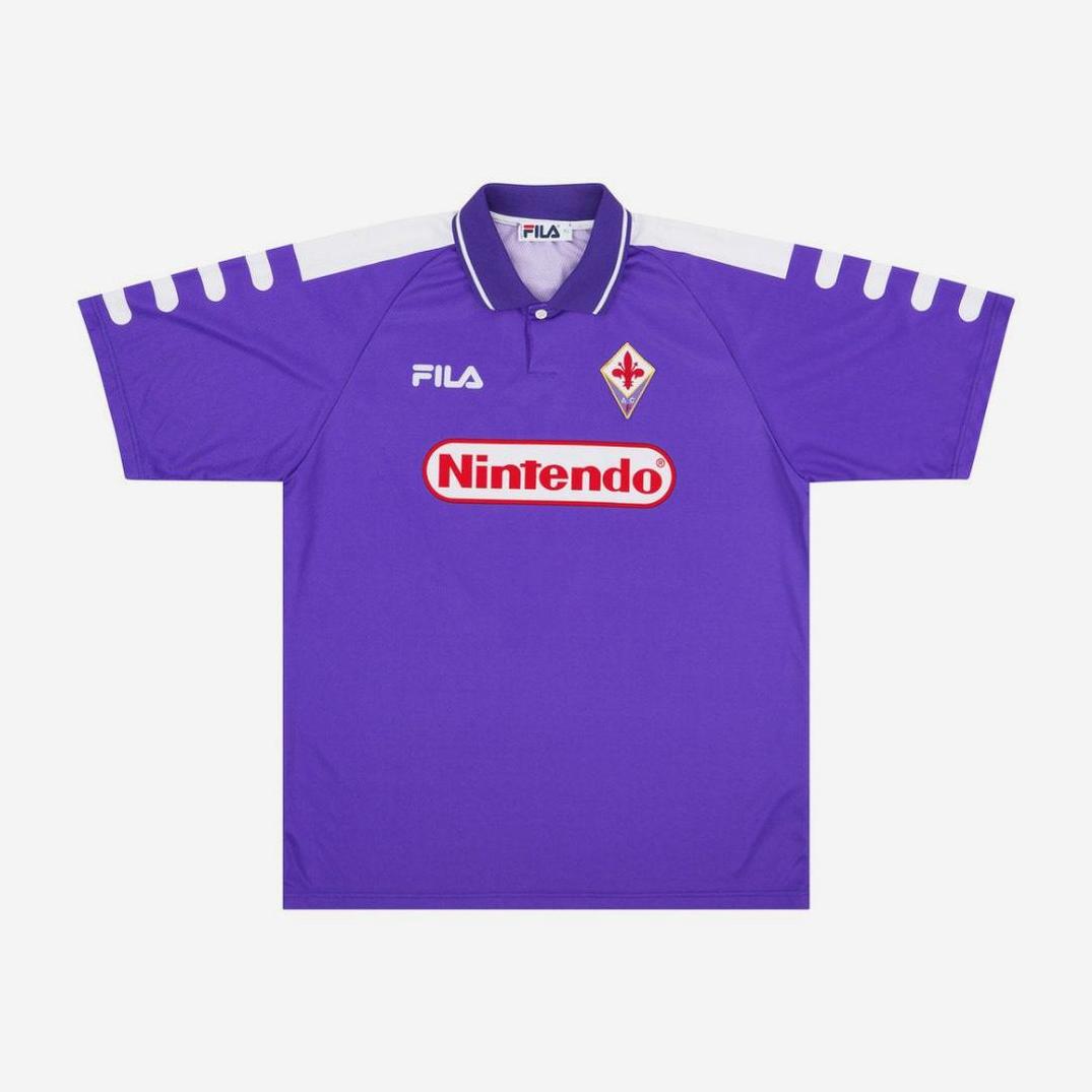 The Retro1999 Fiorentina 1998-99 Home Shirt features a classic purple design with white shoulders, FILA and Nintendo logos, and the club crest—an iconic piece of Serie A history. Displayed here on a plain white background.