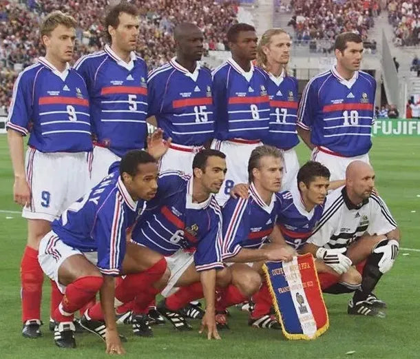 Eleven male soccer players in Retro1999’s France 1998 Home Shirts pose for a pre-match team photo on the field, the front row kneeling and the back row standing, with a cheering stadium crowd in the background.
