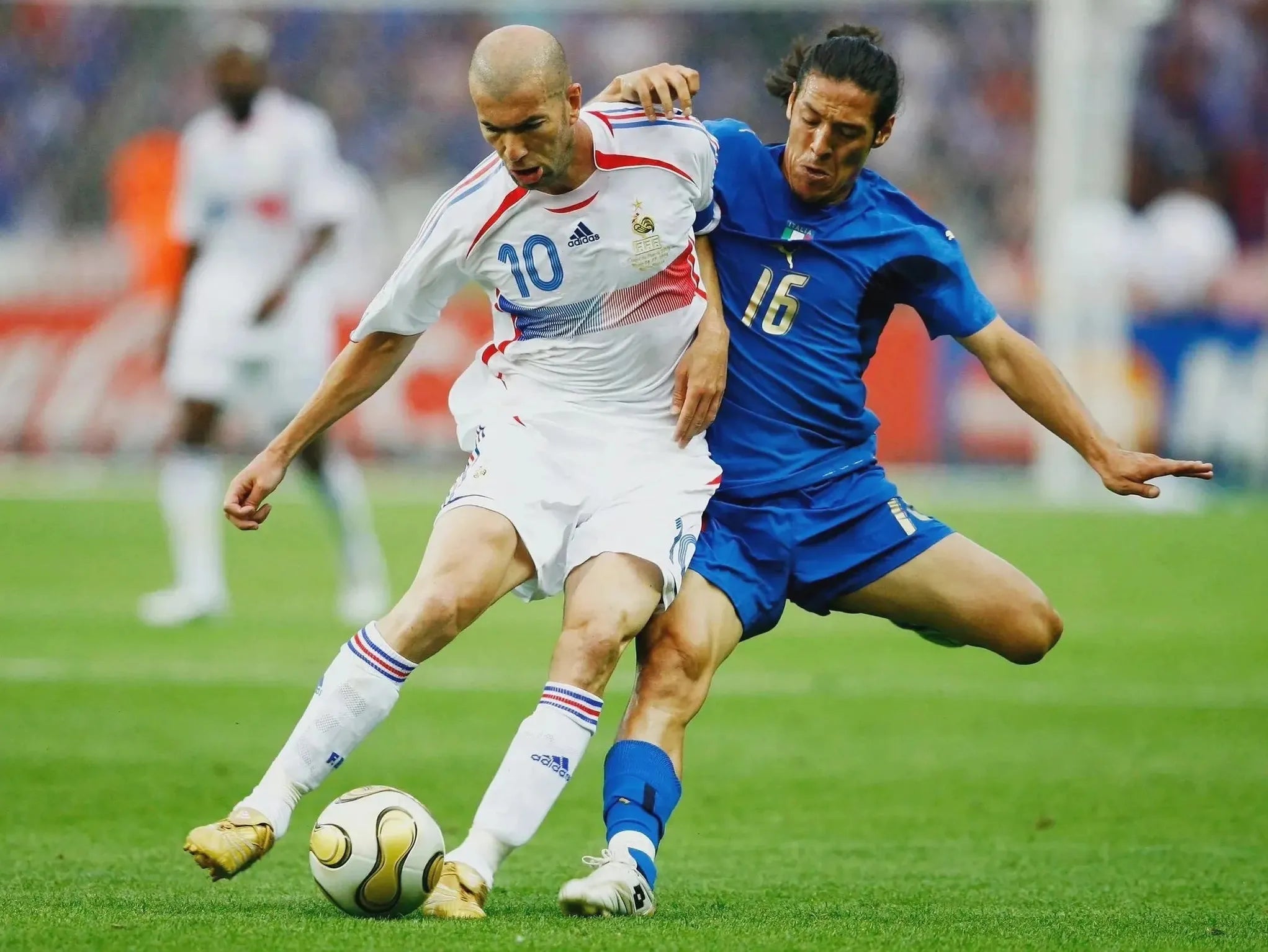A soccer player in a white jersey, number 10, controls the ball as an opponent in a blue Retro1999 France 2006 Away Shirt, number 16, challenges him during the match. Both are focused with a blurred stadium crowd behind them.