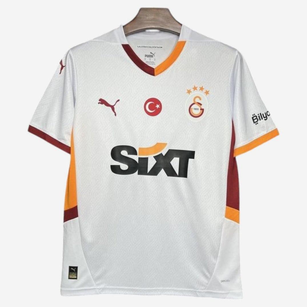 The Galatasaray S.K. 2024-25 Away Shirt by Retro1999 features a white base with orange and dark red accents, Puma logo, Turkish flag, club crest with three stars, and Sixt sponsor logo on the front—proudly displayed on a hanger.