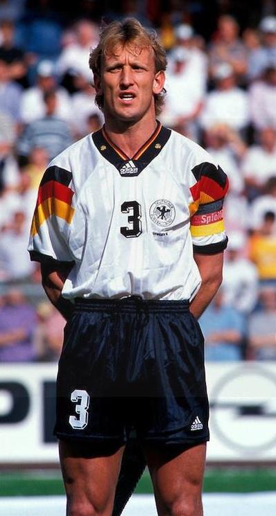 A male soccer player in a Retro1999 Germany 1992 Home Shirt, featuring black, red, and yellow stripes with the number 3, stands on the field hands behind back—a timeless look for football fans.