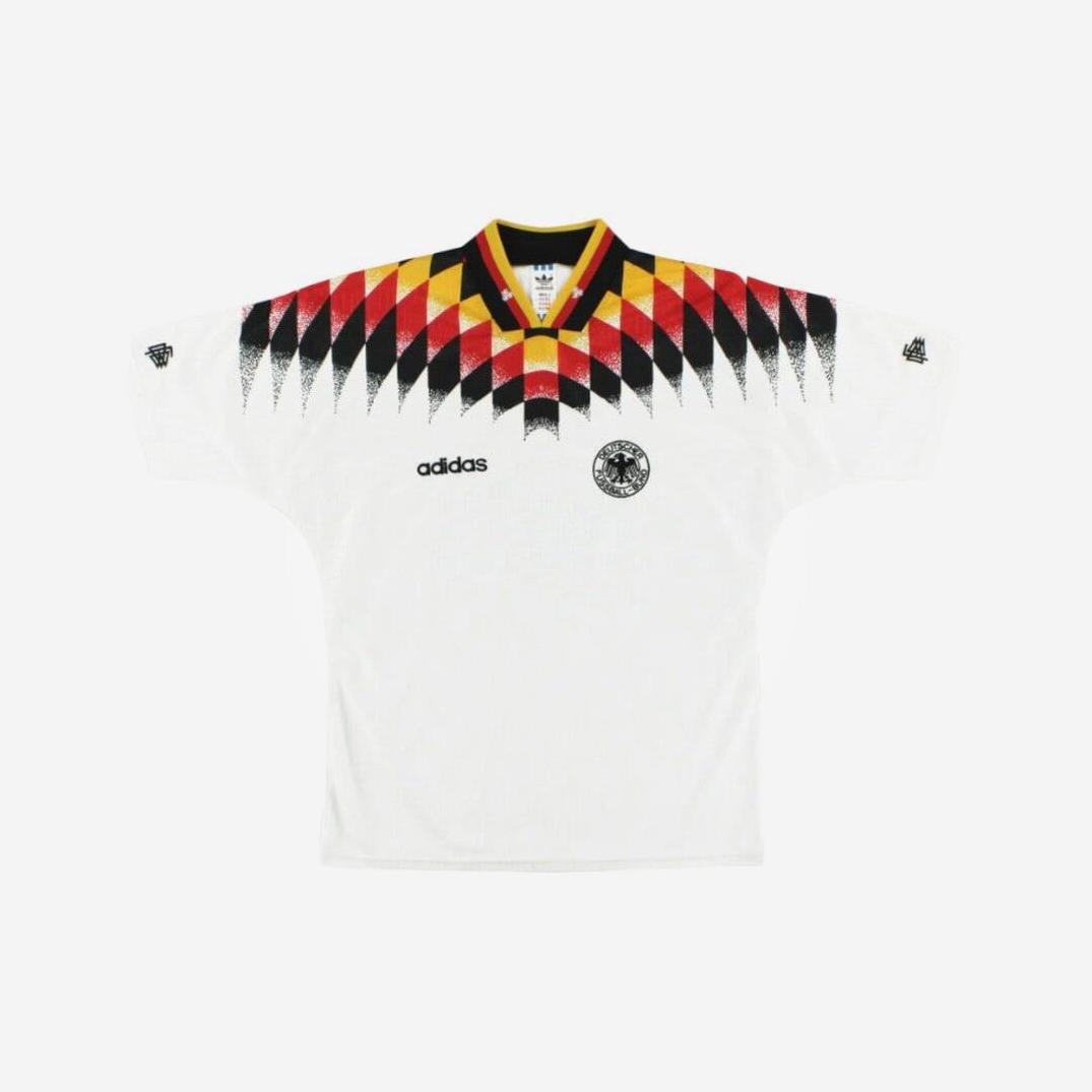 The Germany 1994 Home Shirt by Retro1999 is a vintage white top with bold black, red, and yellow geometric shoulder patterns, plus team logo and brand details—celebrating the German football legacy.