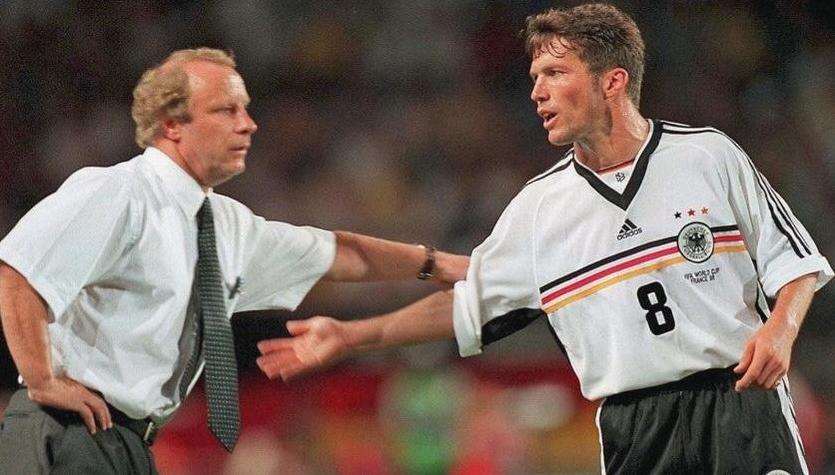 A football coach in a white shirt and tie gestures to a player in the Retro1999 Germany 1998 Home Shirt with number 8. The player, wearing this classic kit, extends his arm toward the coach, capturing a moment of football heritage.