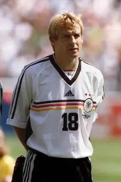 A blonde male soccer player in the Retro1999 Germany 1998 Home Shirt, with black, red, and yellow stripes and number 18, stands on the field with hands behind his back—capturing football history.