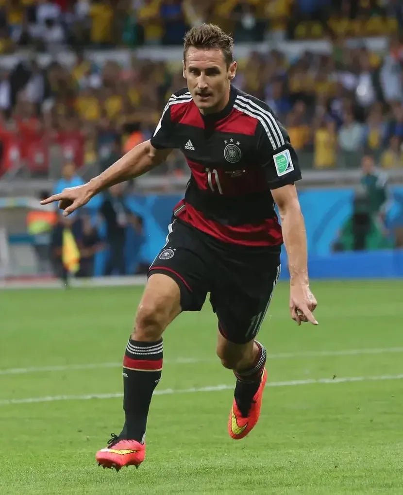 A male soccer player in a Retro1999 Germany 2014 Away Shirt, number 11, runs on the field during a match, with blurred spectators in the background.