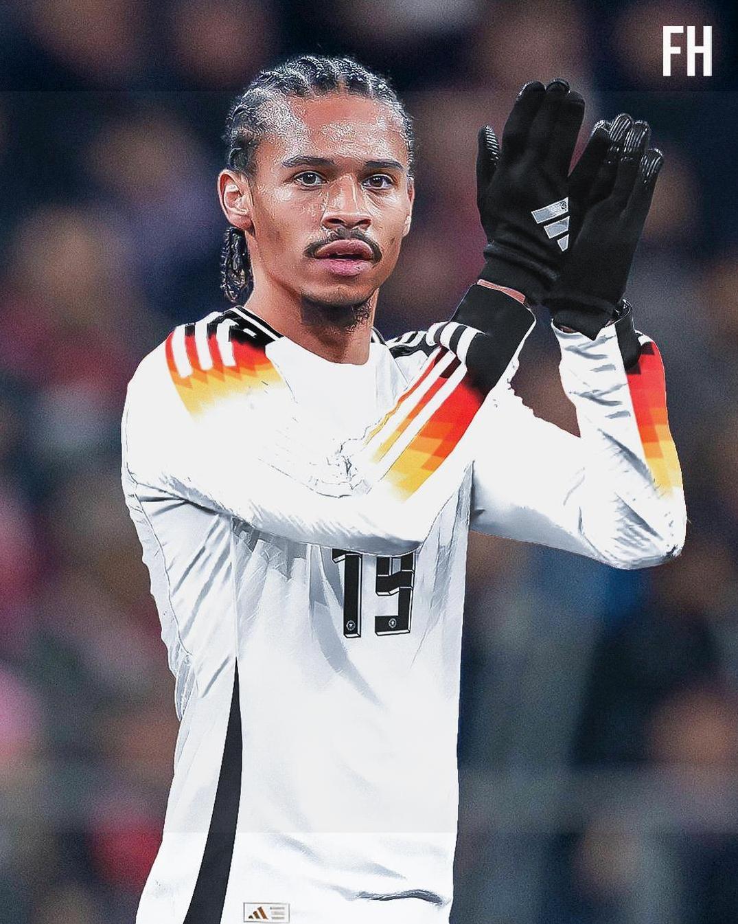 A male Germany national team soccer player wearing the Retro1999 Germany 2024 Home Shirt with number 19 and black gloves claps, his braided hair visible, against a blurred crowd backdrop.