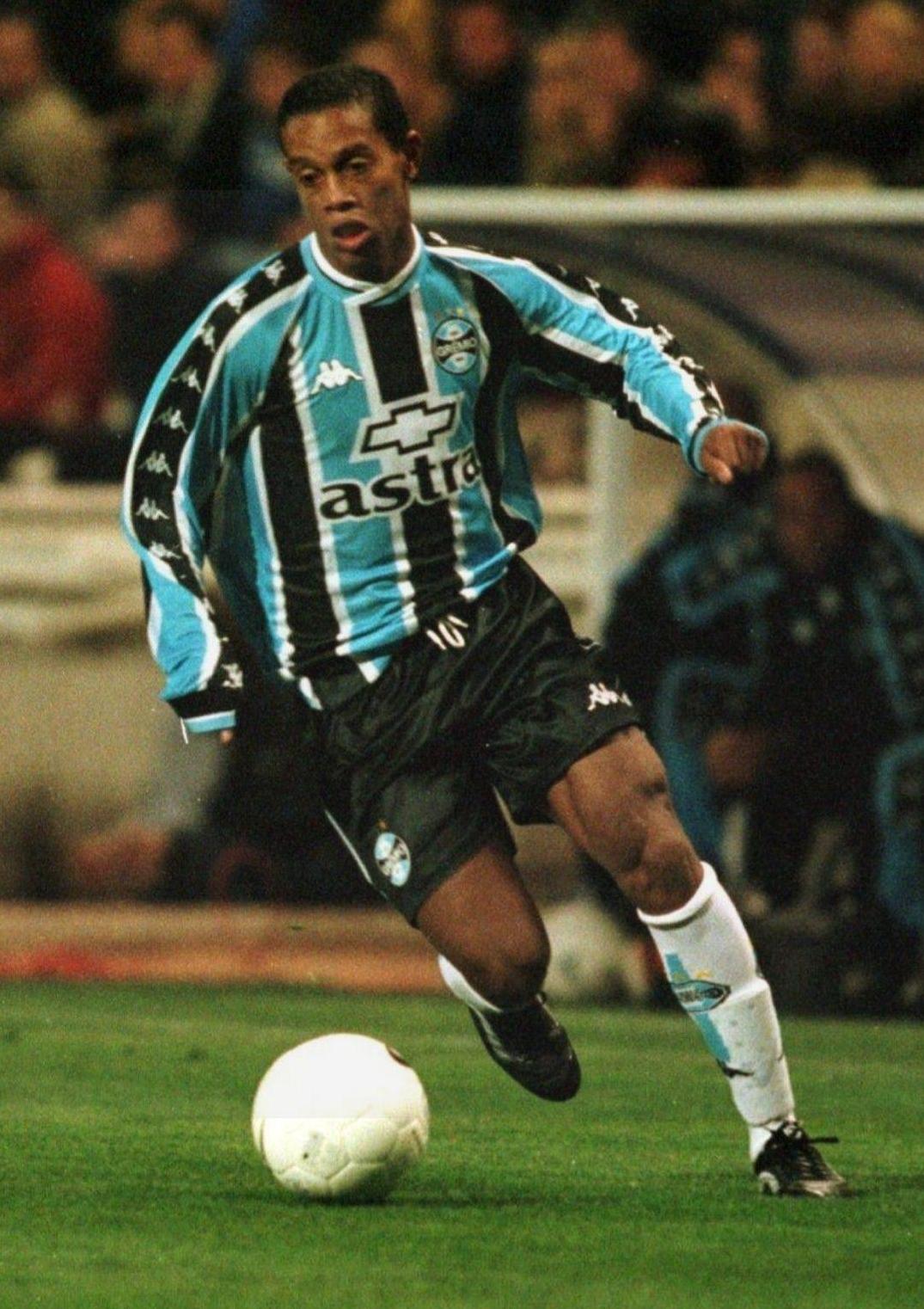 A soccer player sporting the Retro1999 Grêmio 1999-00 Home Shirt dribbles a white ball on the field, paired with black shorts, white socks, and black shoes. Spectators can be seen in the blurred background.