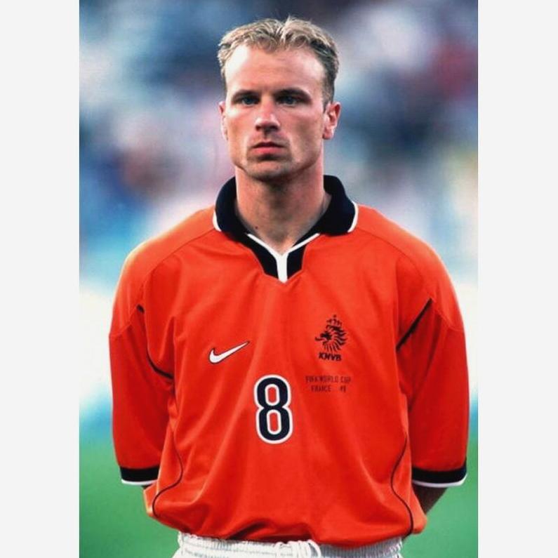 A football player in a Retro1999 Holland 1998 Home Shirt with number 8 stands seriously on the field before a match, the bright orange jersey contrasting with the blurred background.