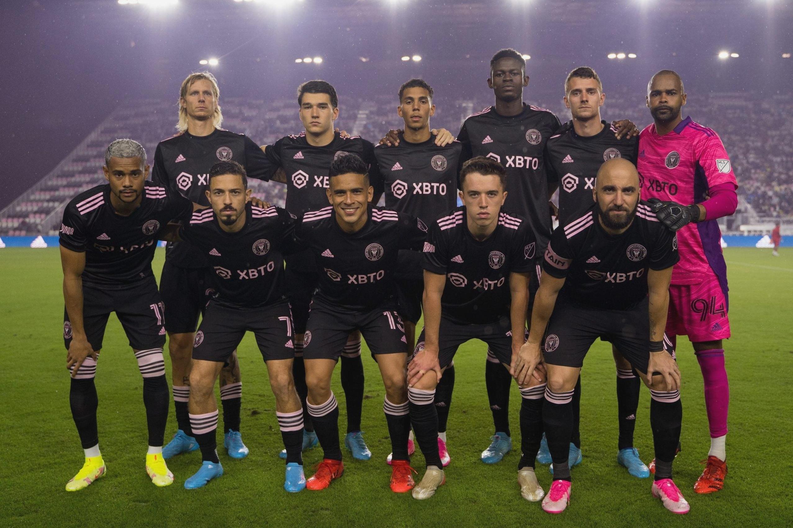 A soccer team wearing Retro1999 Inter Miami 2023/24 Away Shirts with black and pink accents poses for a group photo before an MLS match; their goalkeeper stands to the right in a bright pink kit.