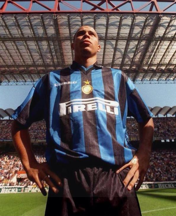 A soccer player in a Retro1999 Inter Milan 1997-98 Home Shirt stands confidently with hands on hips in a stadium, crowd and metal roof visible overhead, recalling memories of Ronaldos iconic debut season.