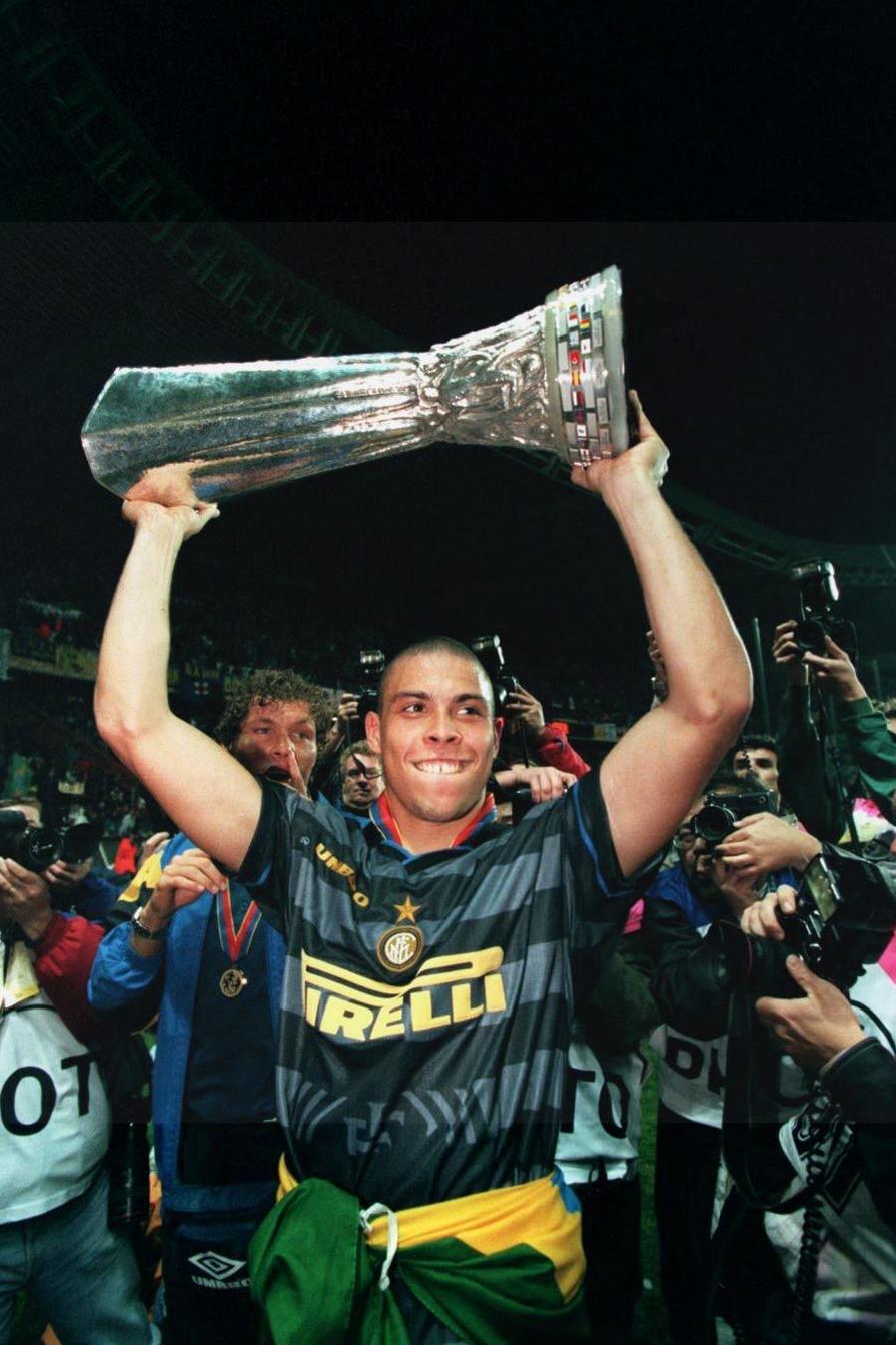 A soccer player in the Retro1999 Inter Milan 1997-98 Third Shirt lifts a trophy above his head, smiling amid photographers and cheering fans on the stadium field.