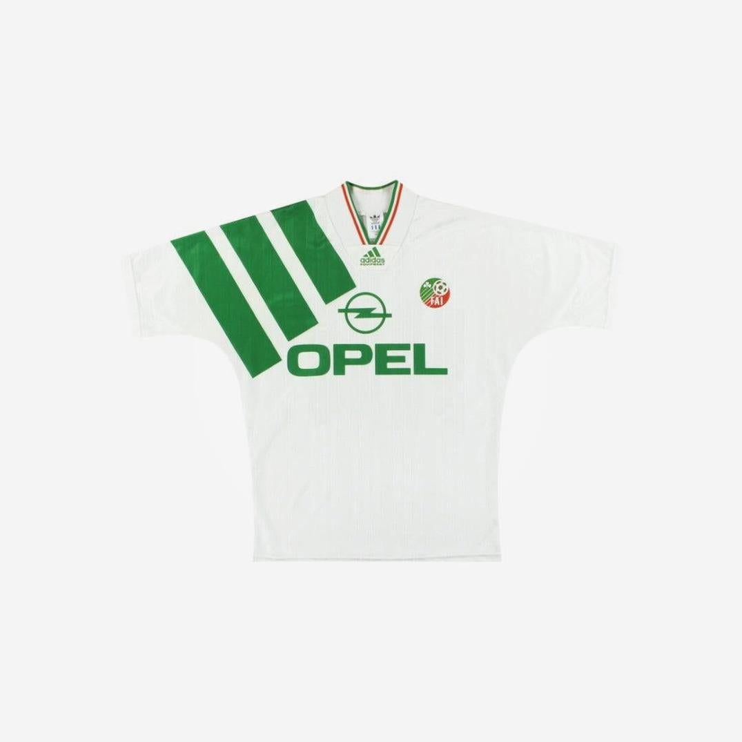 The Retro1999 Ireland 1992/94 Away Shirt is a white soccer jersey with wide green diagonal stripes, Opel and Adidas logos, tri-color collar trim, and a small circular team crest on the left chest—a classic vintage football jersey.