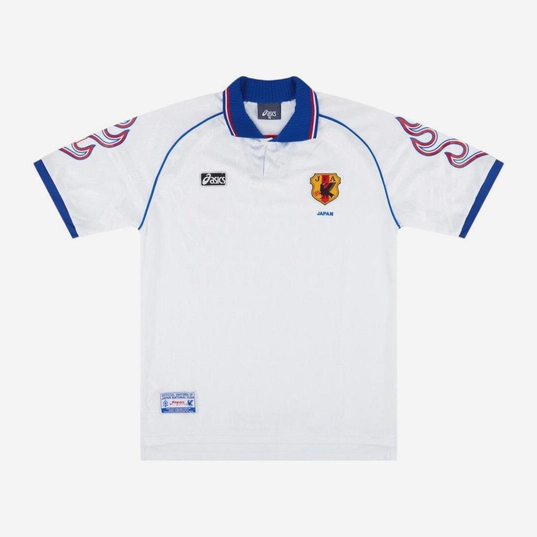 The Japan 1998 Away Shirt by Retro1999 is a white jersey with blue trim, a collar featuring red and blue stripes, a red and yellow crest, and wavy retro designs on the shoulders and sleeves, inspired by Japanese football history.