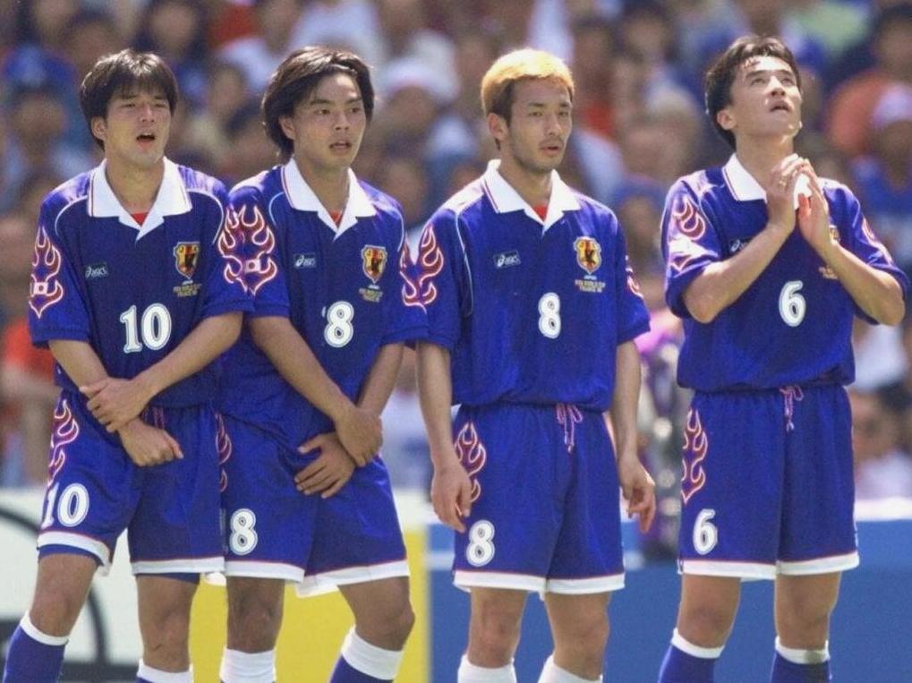 Four male soccer players wear the Japan 1998 Home Shirt by Retro1999, forming a defensive wall during a match. Three protect their bodies with their hands, while the fourth clasps his hands and looks up. A crowd appears in the background.