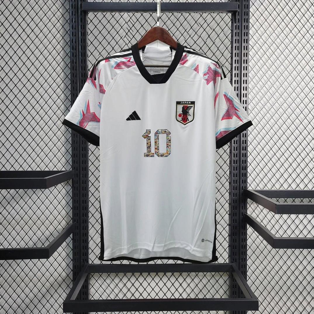 A white Retro1999 Japan 2022 Away Shirt featuring black trim, number 10, team badge, and pink shoulder patterns hangs on a wooden hanger against a mesh wire backdrop.