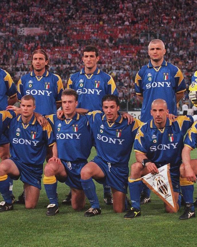 Juventus players wear the Retro1999 Juventus 1995-96 Away Shirt as they pose for a pre-match team photo, standing before a packed stadium—a tribute to their classic Champions League Final kit.
