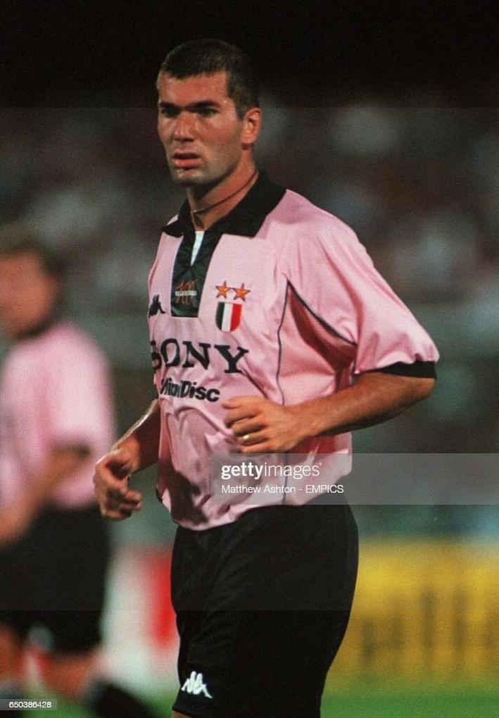 A male soccer player stands on the field in the Retro1999 Juventus 1997-98 Away Shirt with black shorts. The classic kit, featuring the club logo and Sony sponsor, stands out as Serie A champions’ fans blur in the background.