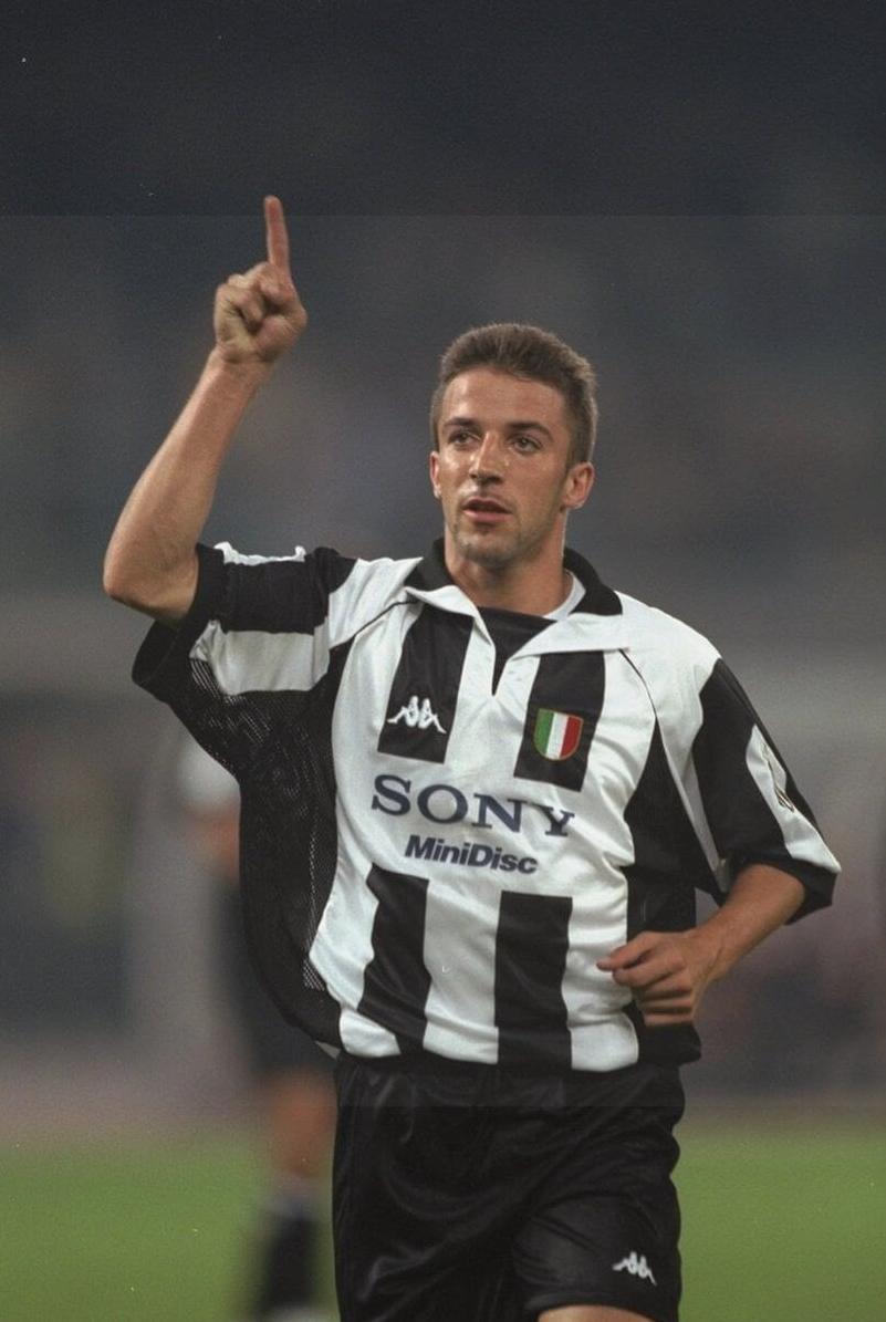 A soccer player in a Retro1999 Juventus 1997-98 Home Shirt raises an arm and points upward, highlighting the authentic design, timeless club heritage, and classic Kappa and Sony MiniDisc logos.