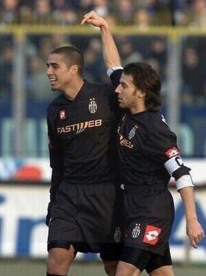 Two soccer players wear the Retro1999 Juventus 2001-02 Away Shirt, celebrating on the field. One smiles with his arm around his focused teammate, arm raised, as spectators watch behind a fence—capturing an authentic retro football moment.