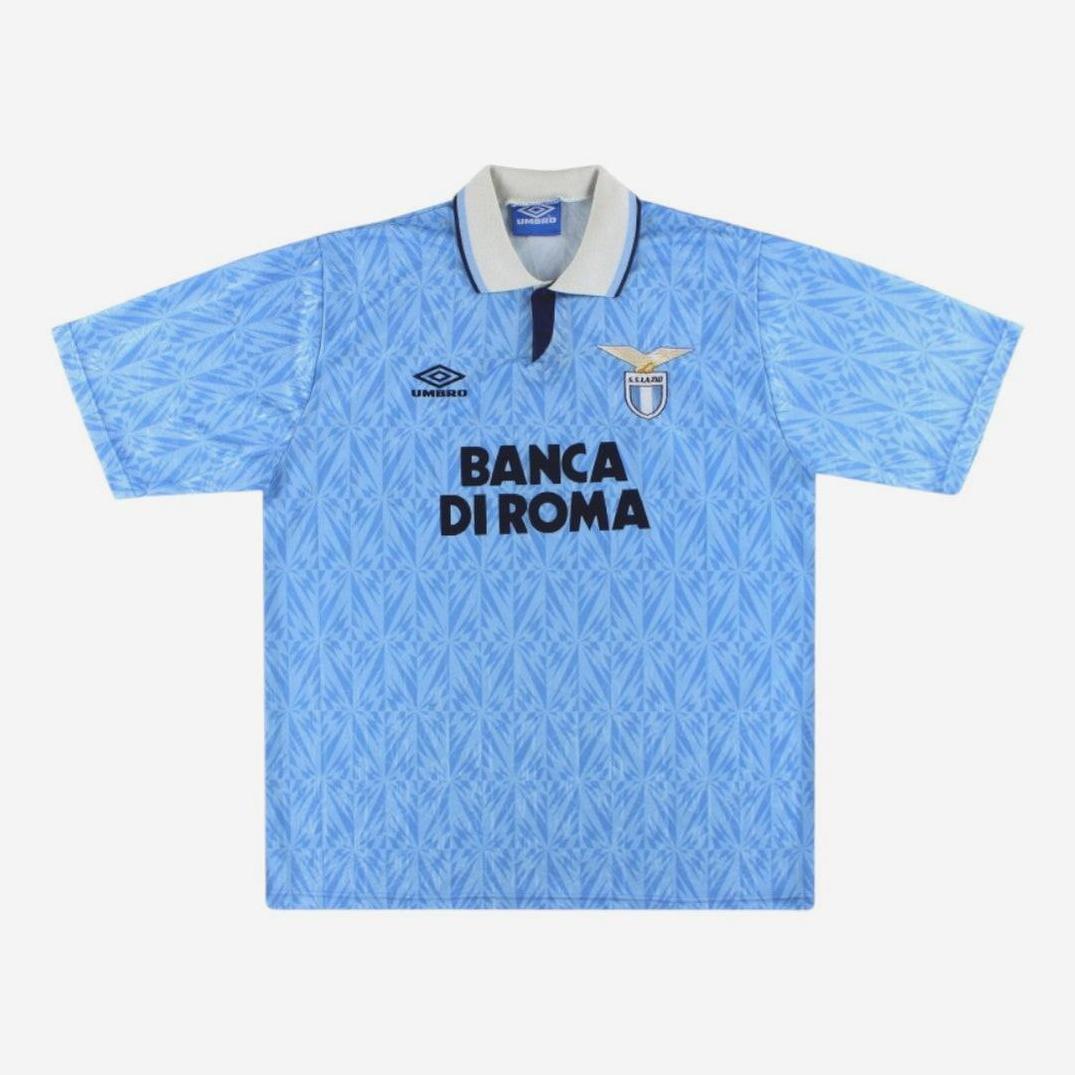 The Lazio 1991-92 Home Shirt by Retro1999 is a light blue football jersey with a geometric pattern, grey collar, black BANCA DI ROMA text, Umbro logo, and classic badge—a must-have retro kit for Lazio fans.