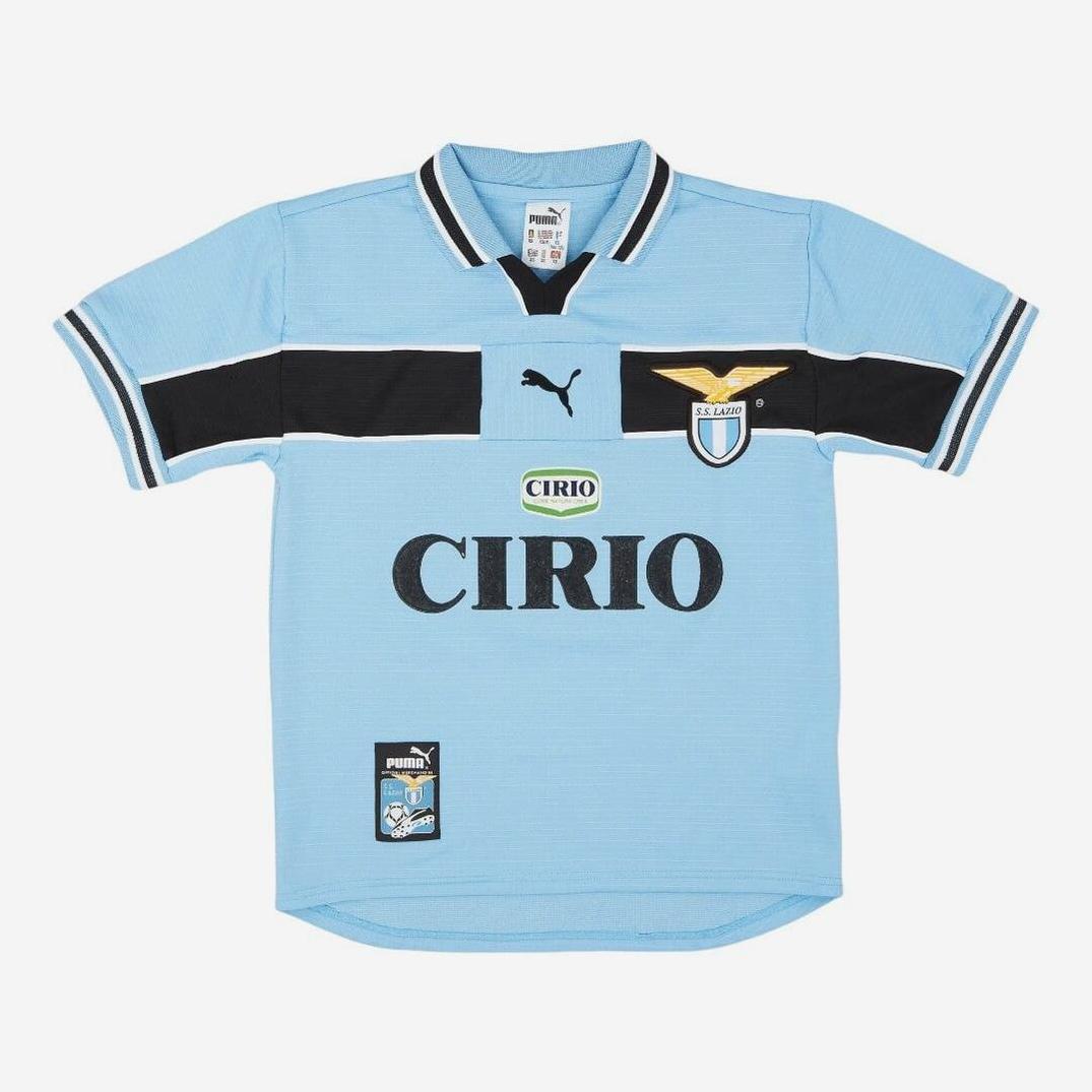 The Lazio 1998-00 Home Shirt by Retro1999 features a light blue design with black and white details, club crest, Cirio sponsor logos, and short sleeves with a collared neckline—a timeless retro choice for Serie A fans.
