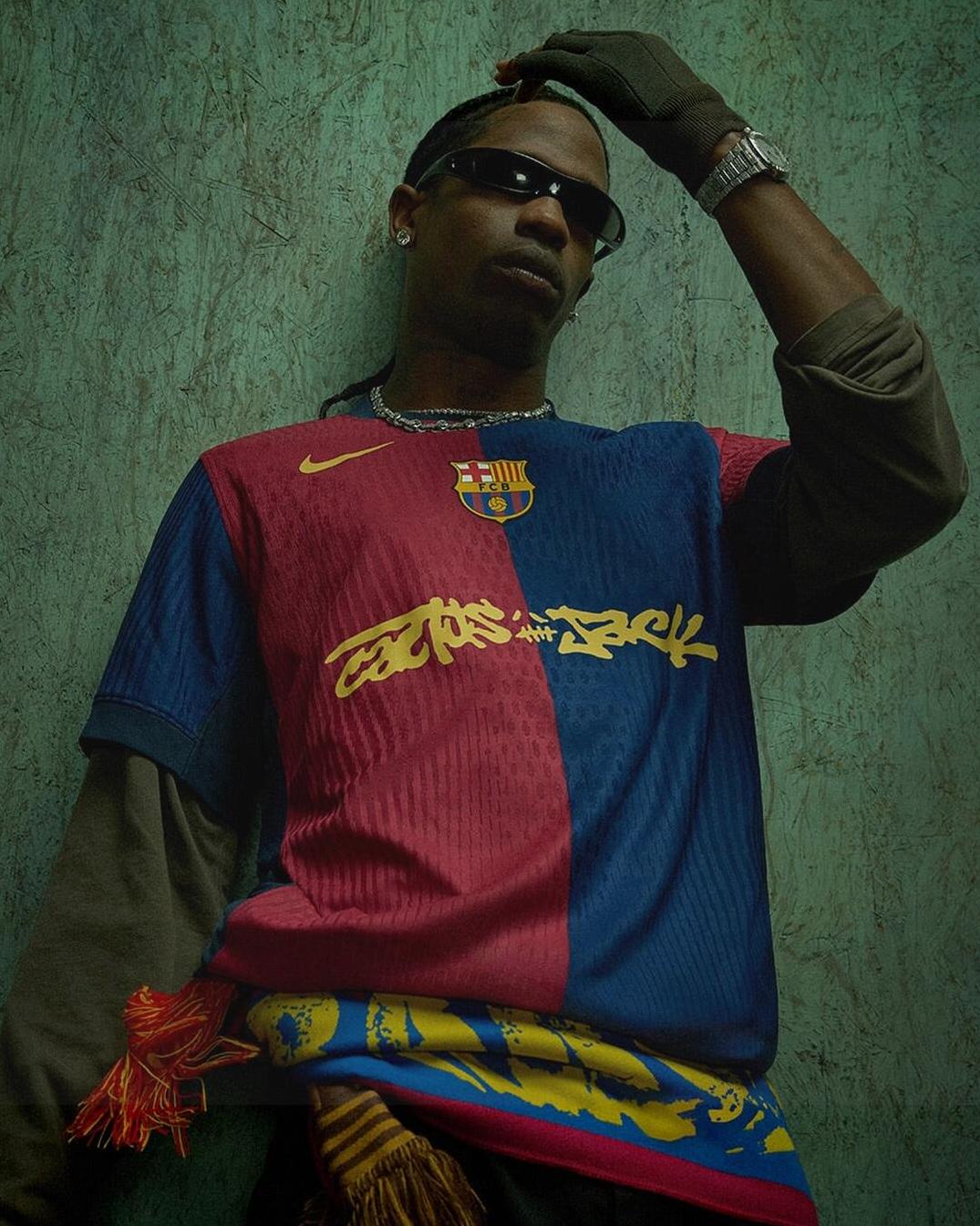 A man in sunglasses and layered clothing poses with one hand on his head, wearing the Limited Edition FC Barcelona X Travis Scott Shirt by Retro1999, featuring graffiti-style writing. He accessorizes with a scarf and watch against a textured wall.