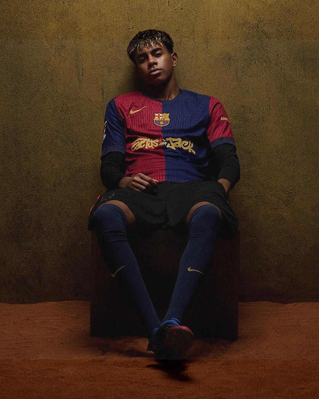 A young male soccer player in the Retro1999 Limited Edition FC Barcelona X Travis Scott Shirt sits on a block by a textured brown wall, arms on legs and feet crossed—a relaxed style for football fans.