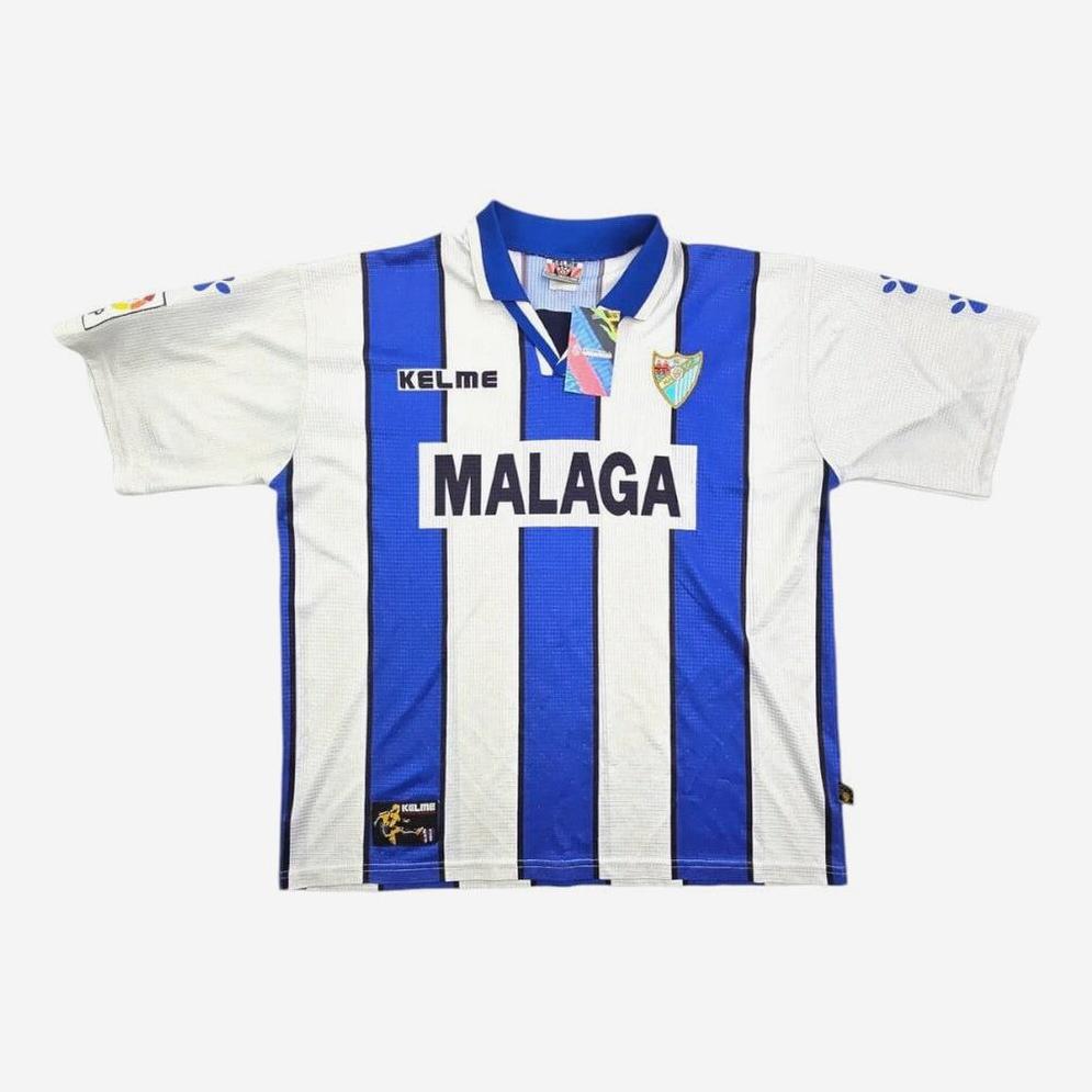 The Malaga 1998-99 Home Shirt by Retro1999 features blue and white stripes, the team crest, bold MALAGA sponsor across the chest, plus blue details on the collar and sleeves—classic style inspired by the original 1998-99 kit.