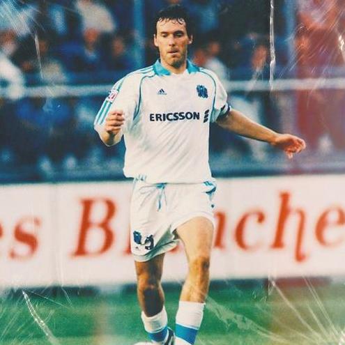A soccer player wears the Retro1999 Marseille 1998-99 Home Shirt with Ericsson logo while running on the field during a match, classic Ligue 1 atmosphere in the background with blurred crowds and advertising boards.