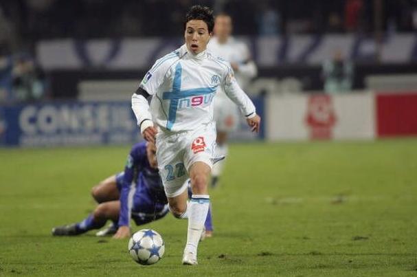 A player in the Retro1999 Marseille 2005-06 Home Shirt sprints with the ball under night lights, while an opponent in a dark kit is on the ground—football fans will appreciate this classic kit on the field.