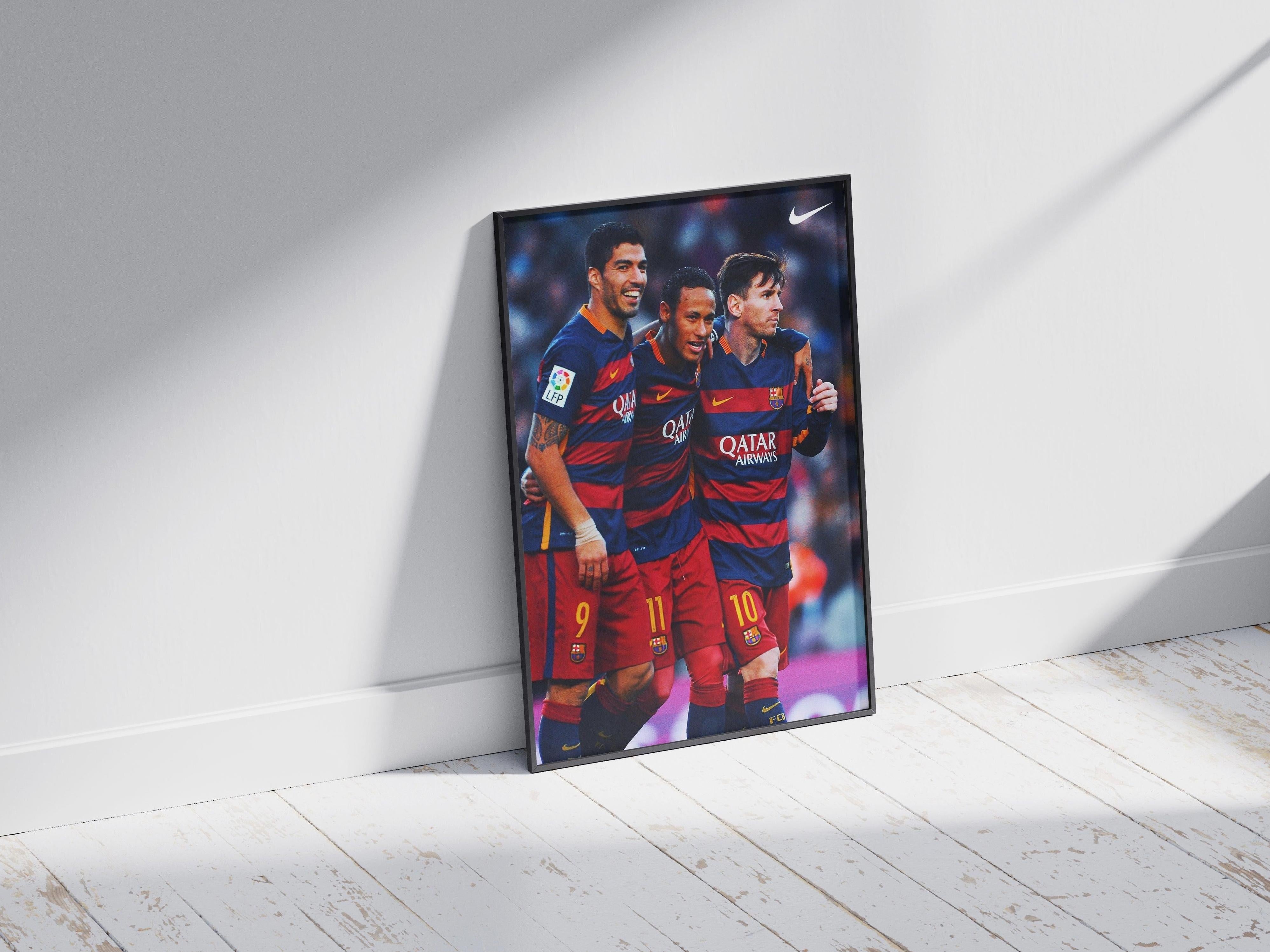 The Retro1999 Messi, Neymar, Suarez MSN Poster features the three FC Barcelona stars in red and blue uniforms, celebrating together on the field, displayed against a white wall on a sunlit wooden floor.