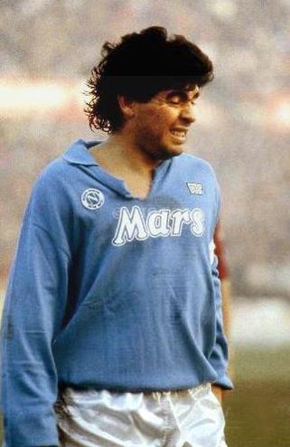 A footballer in a light blue Retro1999 Napoli 1988-89 Home Shirt with Mars sponsor and white shorts stands on the field, squinting, evoking Maradona during the iconic UEFA Cup-winning season.