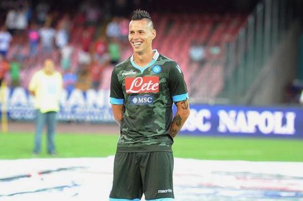 A soccer player in the Retro1999 Napoli 2013-14 Away Shirt stands on the field, smiling with hands behind his back as stadium seats and a blurred crowd highlight Napoli heritage in the background.