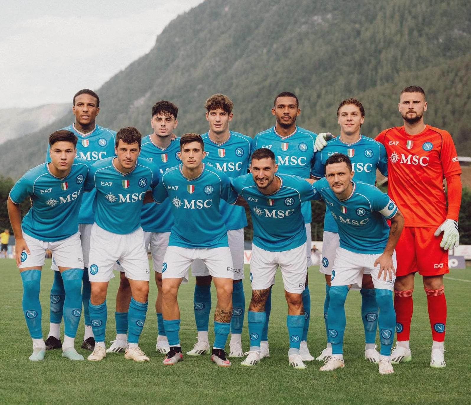 A mens soccer team in blue jerseys and white shorts poses on a grassy field with mountains behind. Some players wear the Retro1999 Napoli 2023-24 Home Shirt, while the goalkeeper stands right in an orange kit.