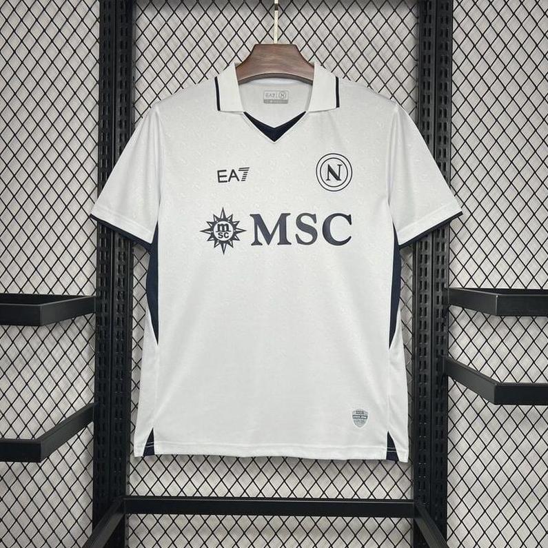 A white Napoli 2024-25 Away Shirt by Retro1999 with black trim hangs on a wooden hanger against a mesh backdrop, showcasing EA7, N, sun emblem, and MSC logos for a classic Serie A club look.