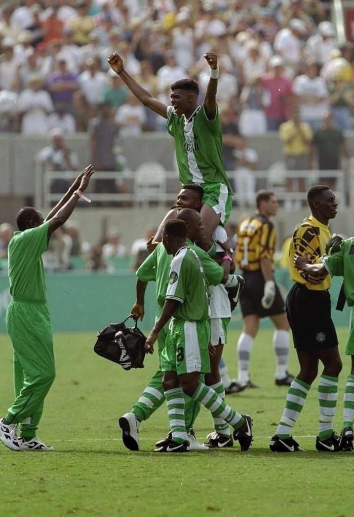 Nigerian soccer players celebrate on the field in the Retro1999 Nigeria 1996 Home Shirt, one Super Eagles star lifted high as teammates applaud, with a yellow-clad goalkeeper and cheering crowd during the African Cup of Nations.