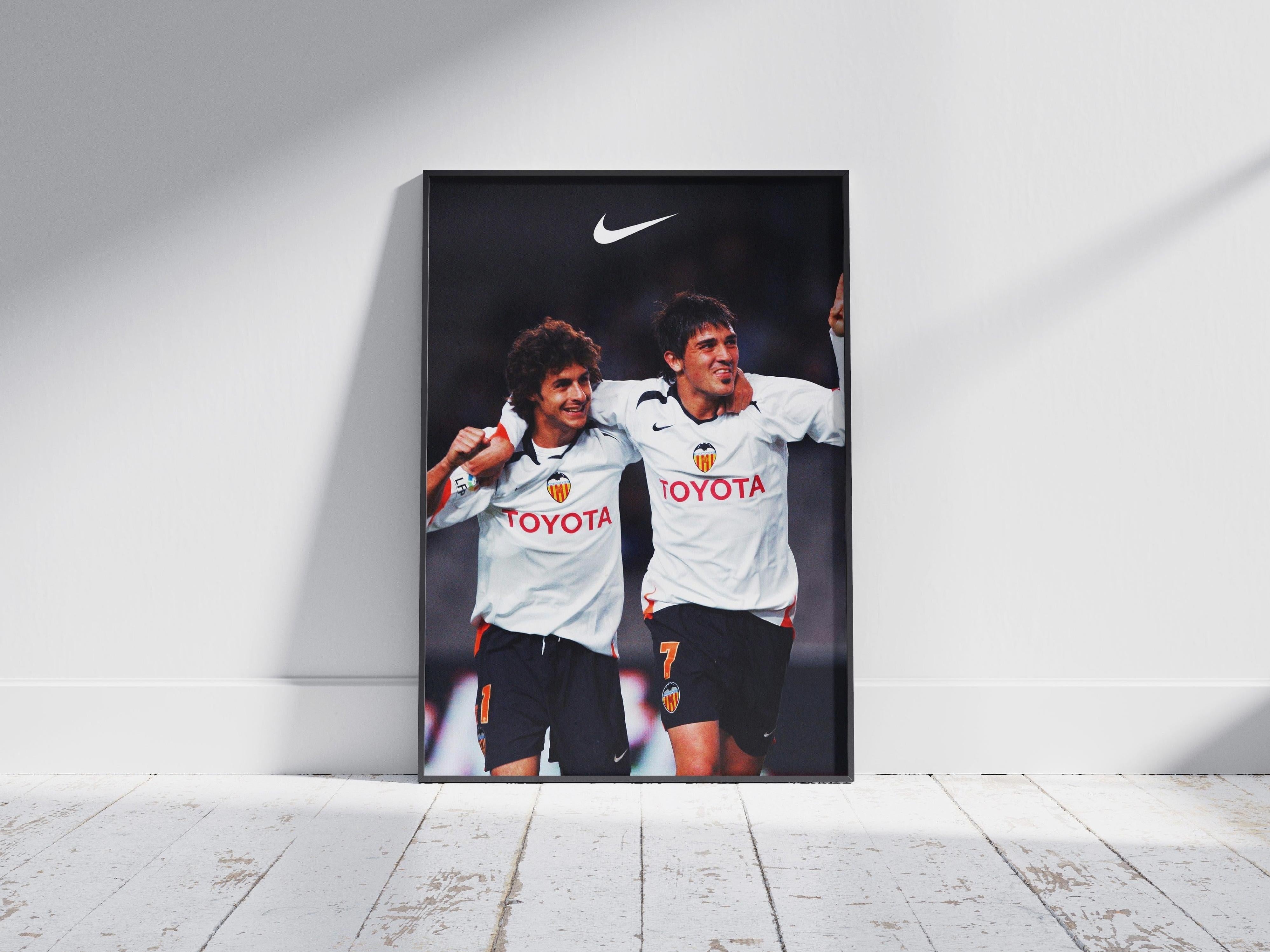 The Pablo Aimar x David Villa Poster by Retro1999 features two Valencia CF stars in classic white jerseys with Toyota sponsor, celebrating passionately. Displayed in a frame against a white wall and light wood floor—ideal Spanish football memorabilia.