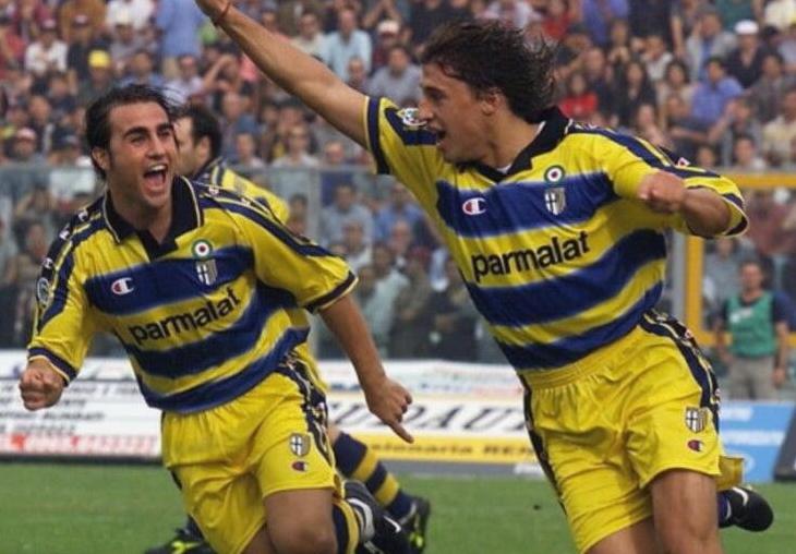 Two soccer players in yellow and blue Retro1999 Parma 1999-00 Home Shirts celebrate on the field, one with his fist raised, as fans cheer them on and the Serie A club revels in classic kit style.