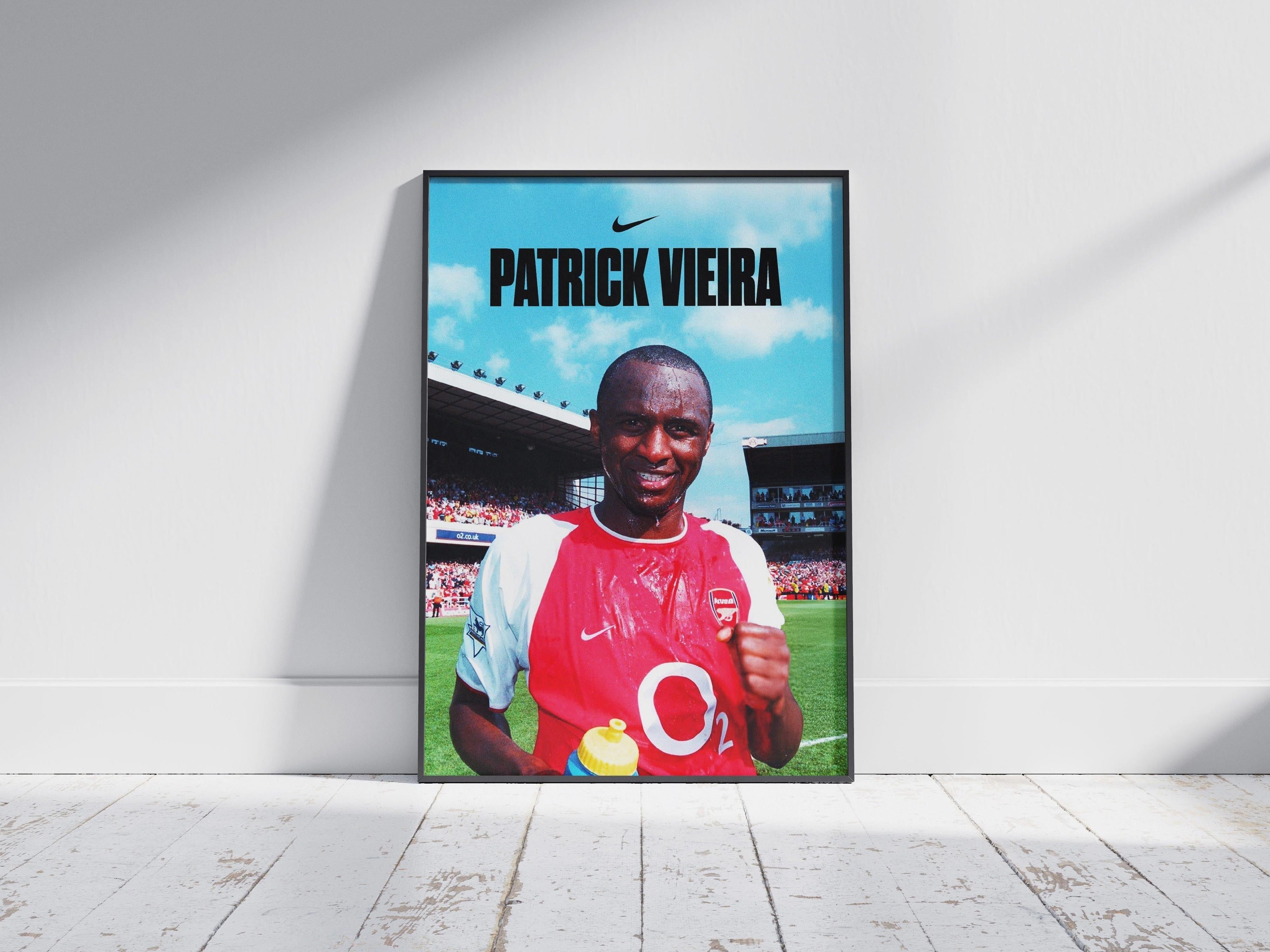 The Patrick Vieira x Arsenal Poster by Retro1999 shows Premier League legend Patrick Vieira in an Arsenal jersey, smiling and holding a drink on the field, with his name and the Nike logo above, framed and set on a wooden floor.