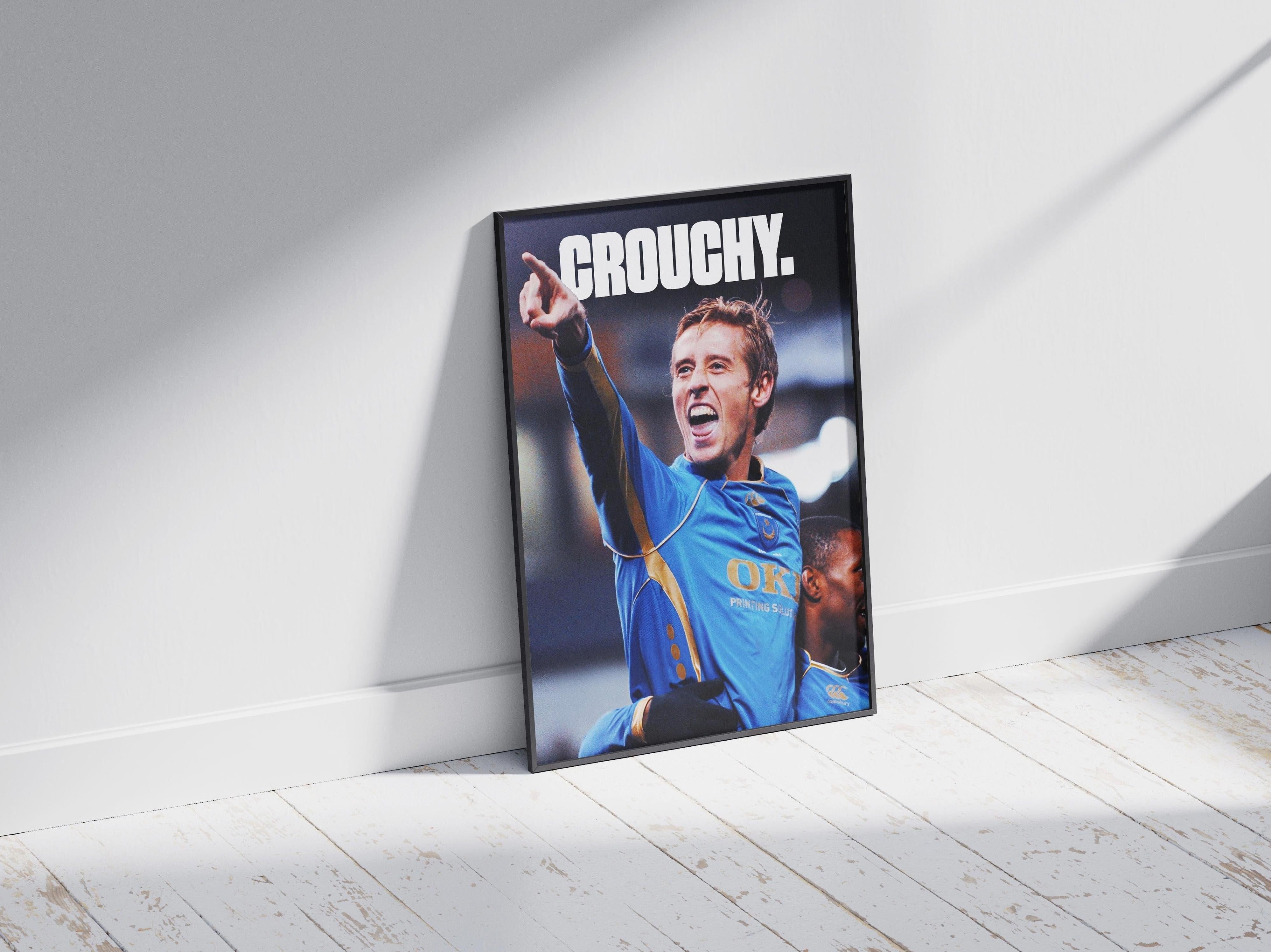 The Peter Crouch x Jermain Defoe Poster by Retro1999 leans against a white wall, featuring a soccer star in a blue and yellow kit with “CROUCHY.” at the top—a nostalgic tribute to EPL legends. Sunlight casts shadows on the wooden floor.