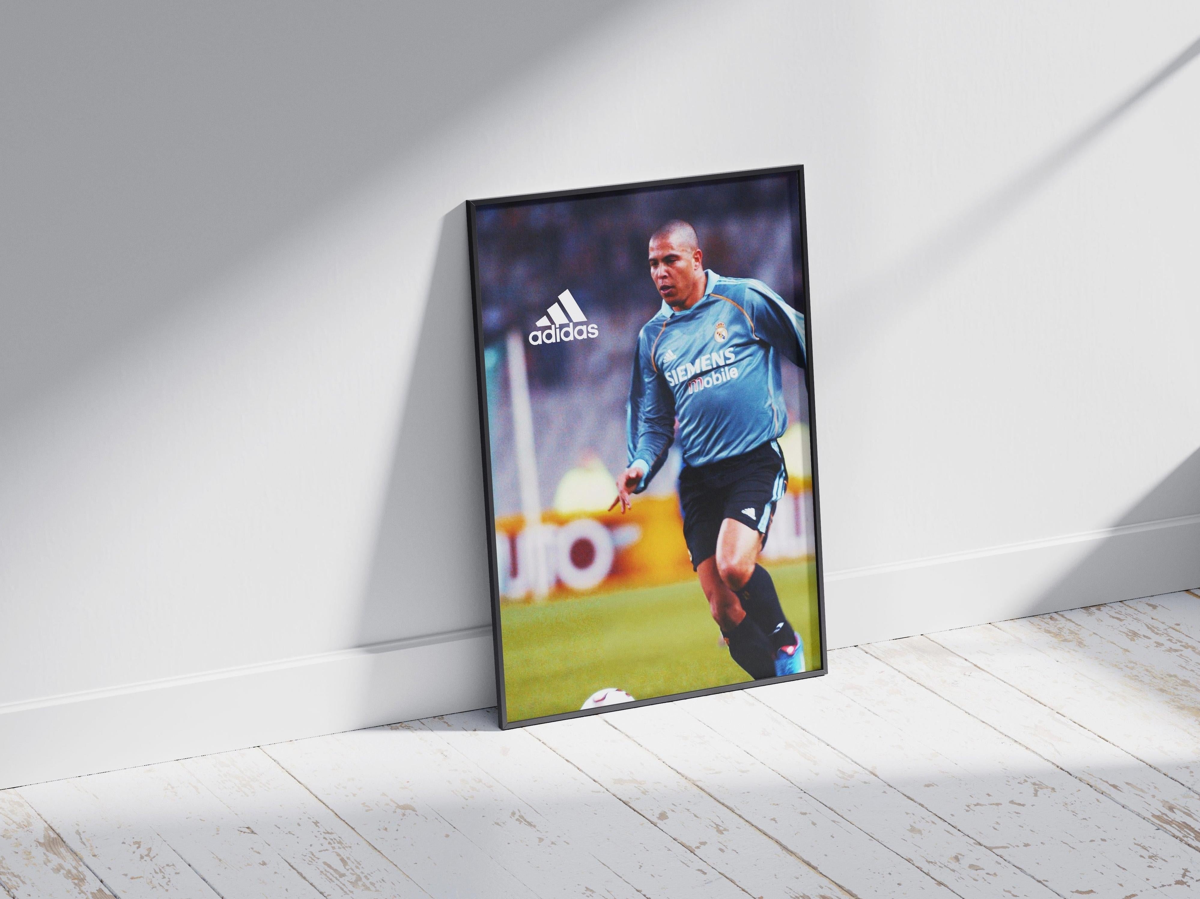 The Retro1999 R9 x Real Madrid Poster shows Ronaldo in a blue jersey and black shorts, framed and displayed against a white wall with sunlight casting shadows—perfect for football fans.