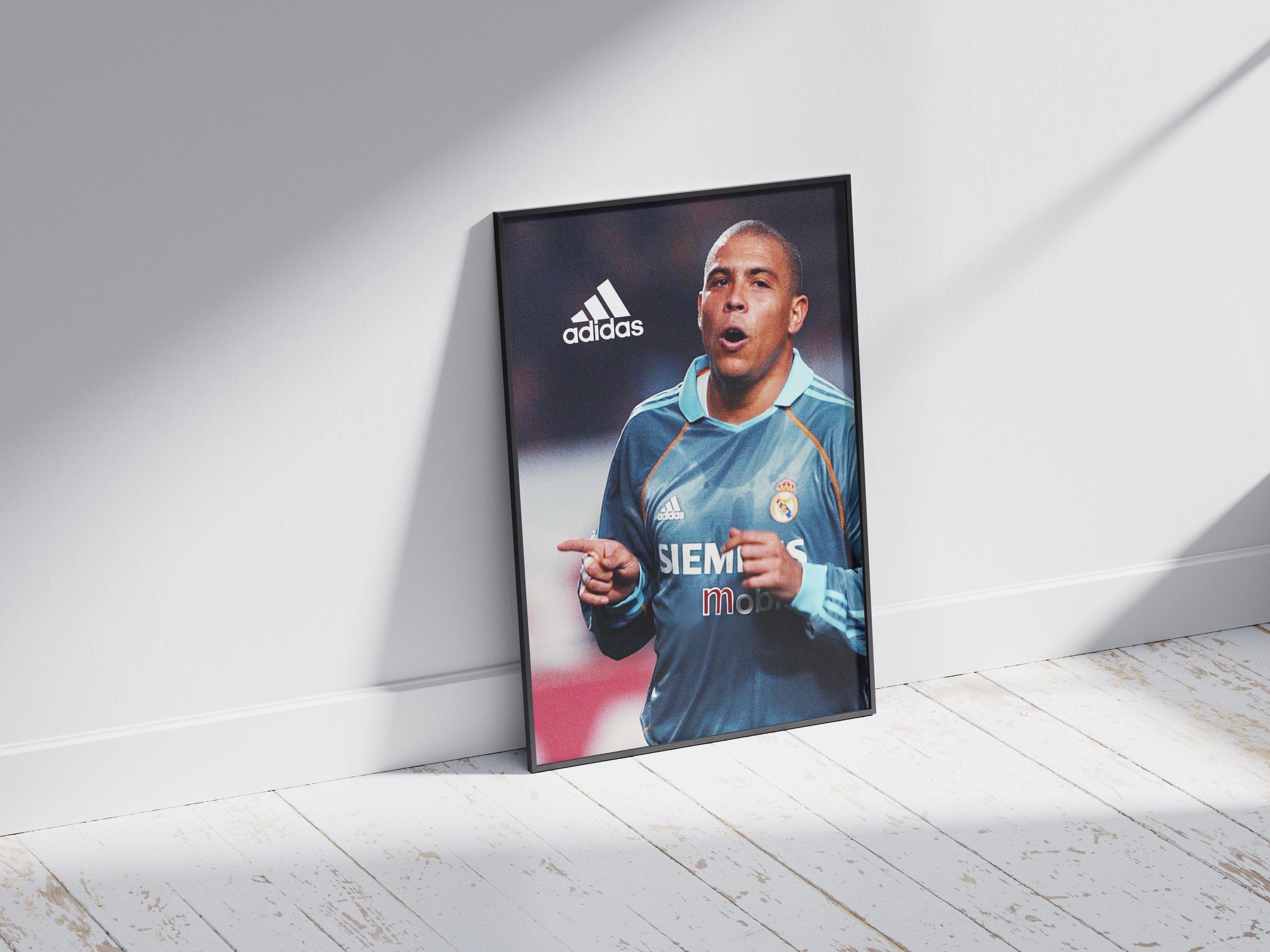 The Retro1999 R9 x Real Madrid Poster shows a soccer player in a blue Adidas/Siemens Mobile kit celebrating with raised hands, linked to the Spanish Primera Liga era, framed and placed against a white wall on a wooden floor.