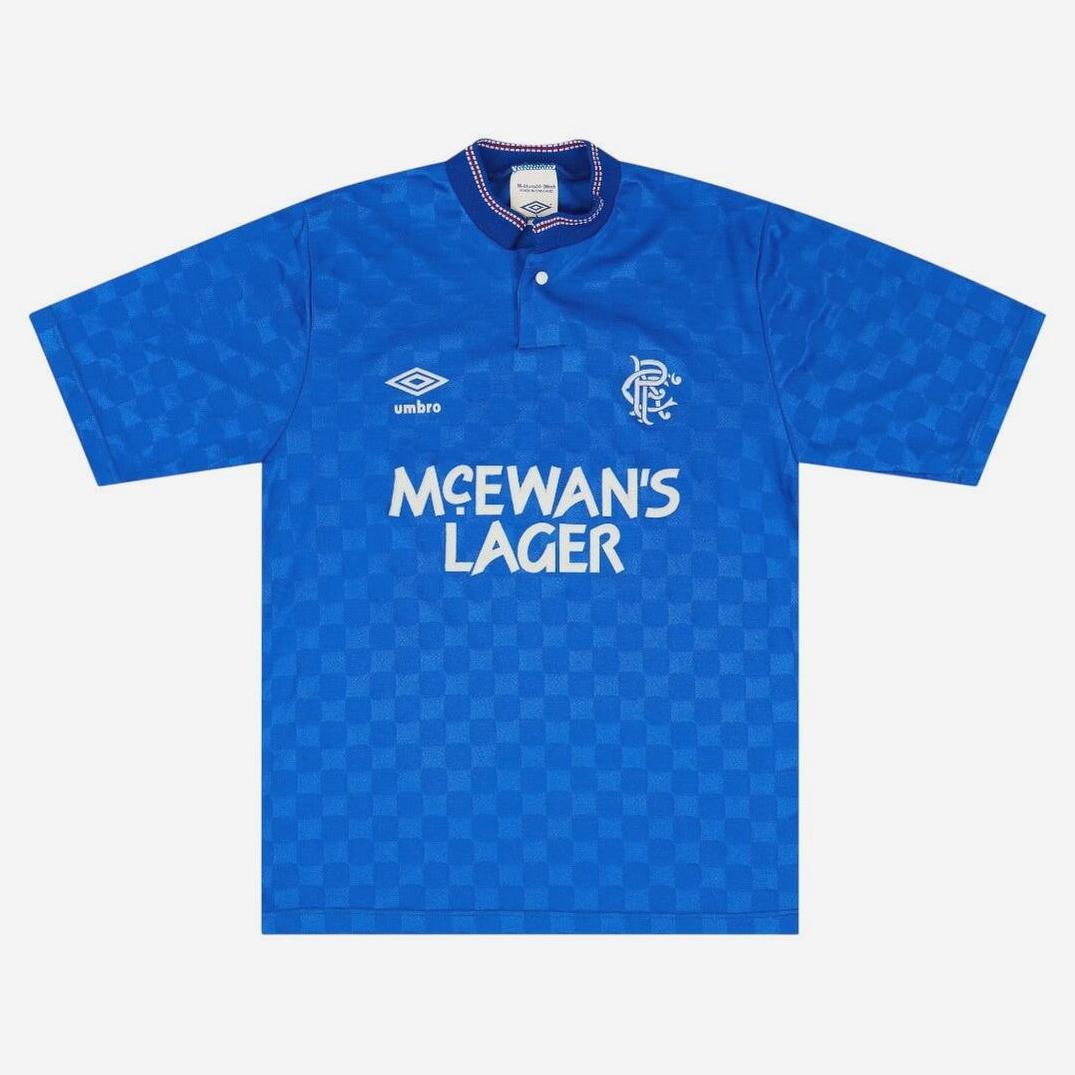 The Retro1999 Rangers F.C. 1987-90 Home Shirt features a blue checkerboard design, short sleeves, buttoned collar with red, white & blue trim, plus McEwan’s Lager sponsor, Umbro logo, and Rangers FC crest on the chest.