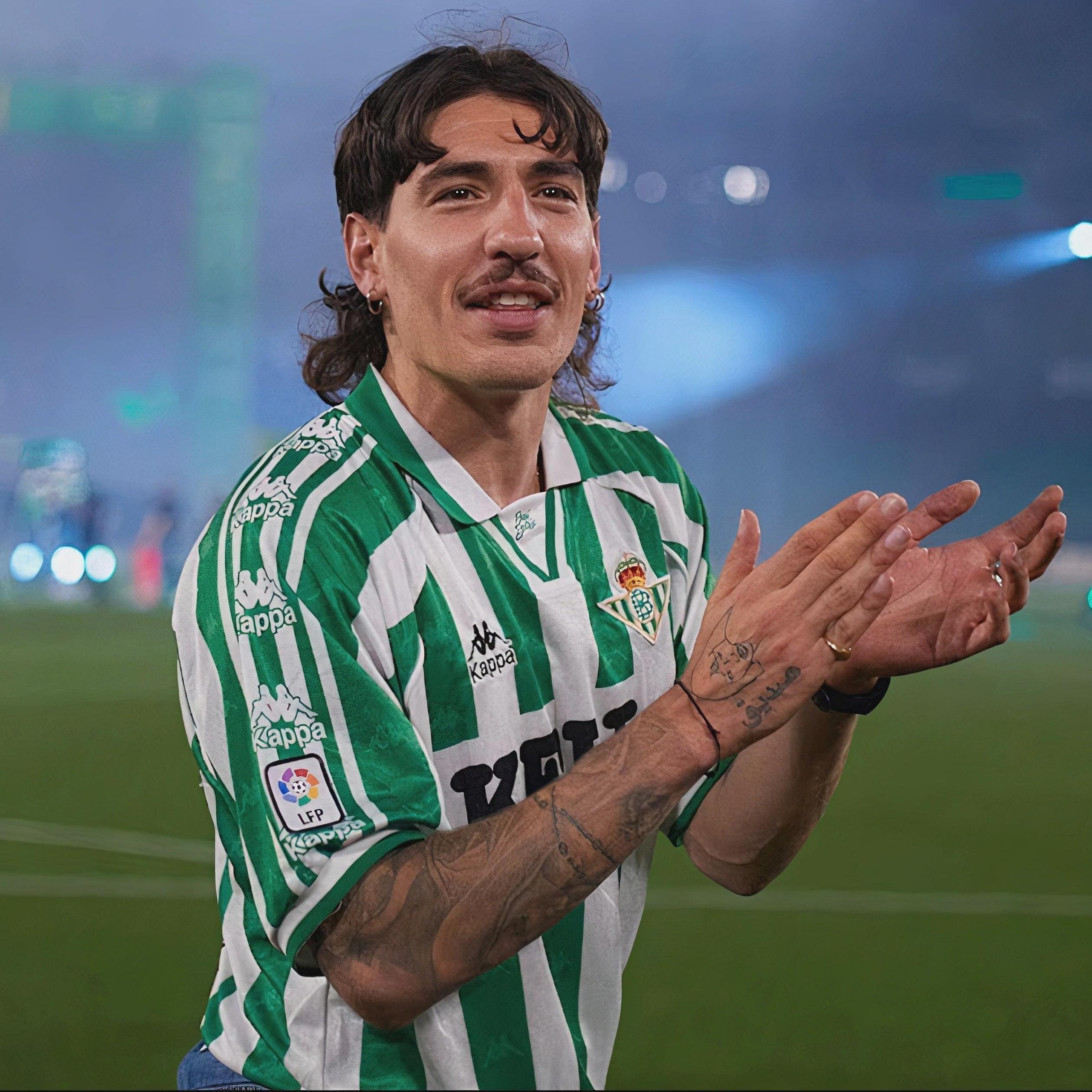 A male soccer player with long dark hair, mustache, and arm tattoos claps on a La Liga field under stadium lights, wearing the Retro1999 Real Betis 1995-98 Home Shirt in green and white stripes.