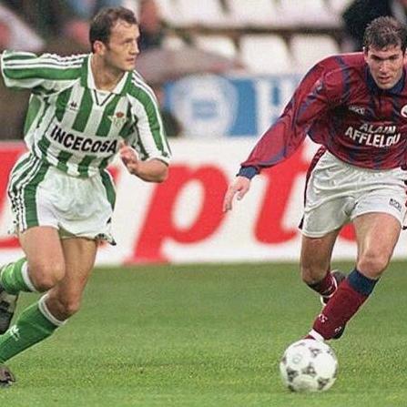 Two soccer players in action: one wearing the Retro1999 Real Betis 1995-98 Home Shirt, the other in red and blue, both chasing a white soccer ball during an exciting La Liga match.
