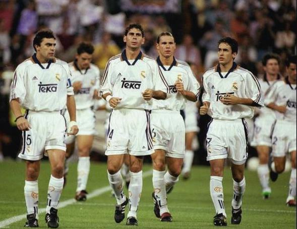 Four Real Madrid players in white uniforms wear the Retro1999 Real Madrid 1999-00 Home Shirt, featuring an authentic retro design, as they walk together on the field during a match with a blurred crowd in the background.