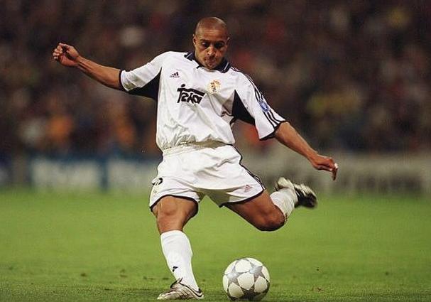 A soccer player wears the Retro1999 Real Madrid 2000-01 Home Shirt while kicking a ball on the field during a La Liga match, with a blurred crowd in the background.
