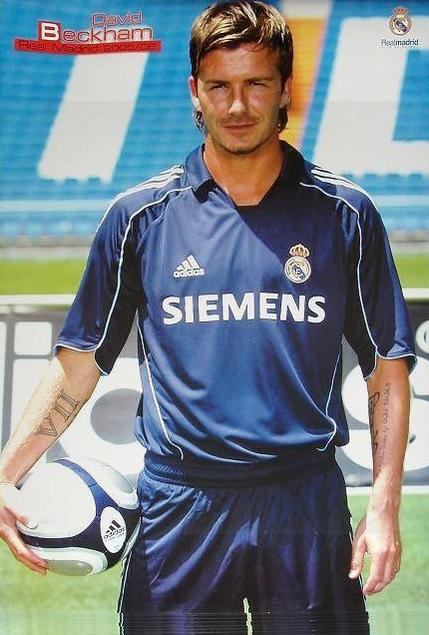 A soccer player in a dark blue Real Madrid 2005-06 Away Shirt by Retro1999 holds a ball under his arm on the field. With short styled hair and tattoos, he highlights this authentic retro collectors item.