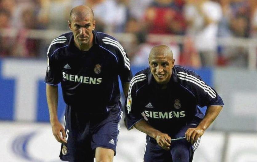 Two soccer players in Retro1999’s Real Madrid 2005-06 Away Shirts, featuring the SIEMENS logo, stand on the field looking ahead, with a blurred crowd in the background.