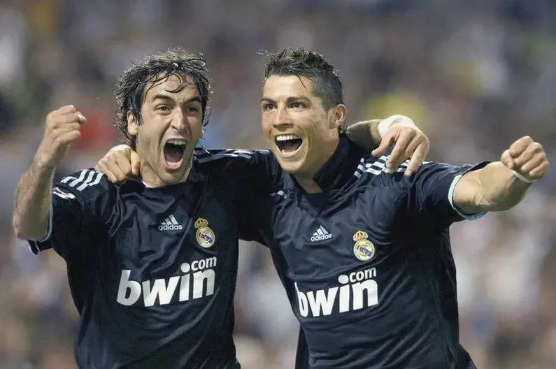 Two soccer players jubilantly celebrate in Retro1999 Real Madrid 2009-10 Away Shirts, arms outstretched and smiles wide, with a blurred crowd behind—a perfect scene for Real Madrid fans.